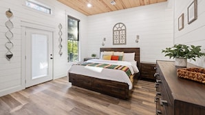 A bedroom with a wooden ceiling, a large bed with a colorful blanket, a wooden dresser, and a window with a view of greenery. - Rustic Hillside Hideaway-2 BR+Bunk-Timber Creek (Broken Bow)