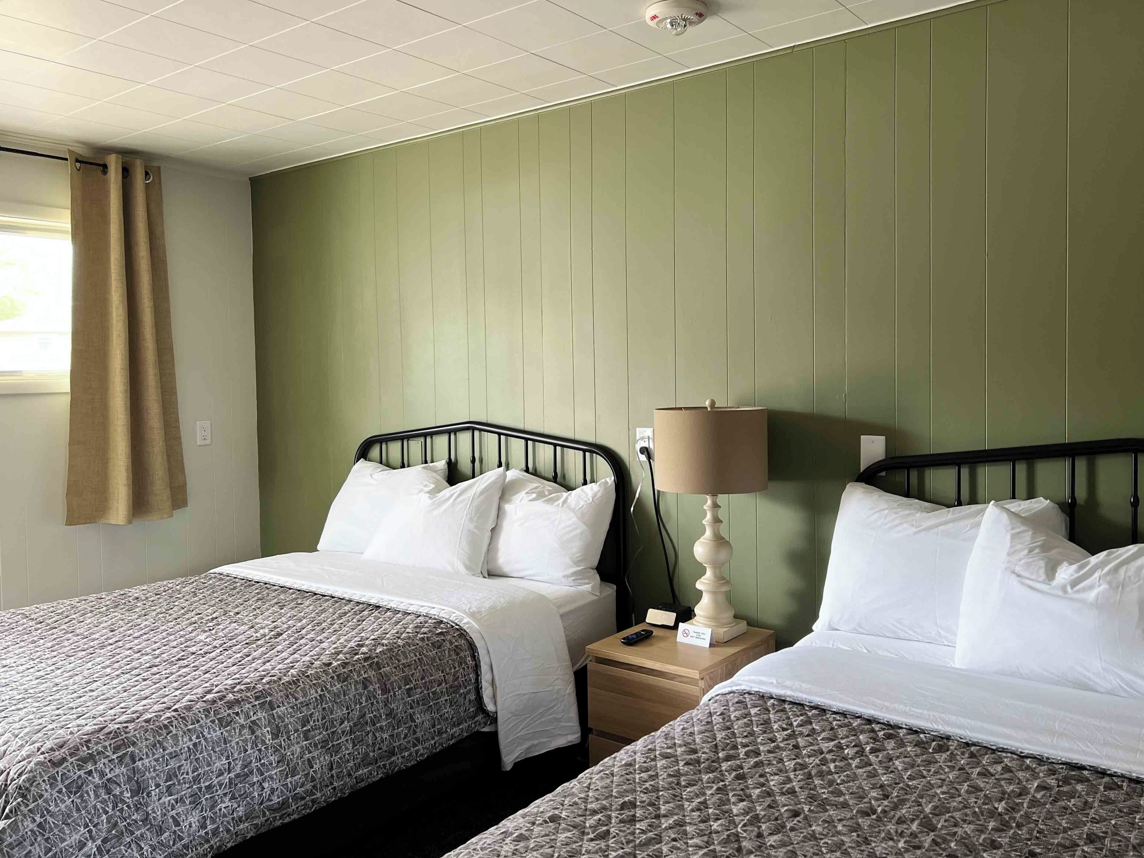 A hotel room with two beds, a nightstand, a lamp, and a window with curtains.