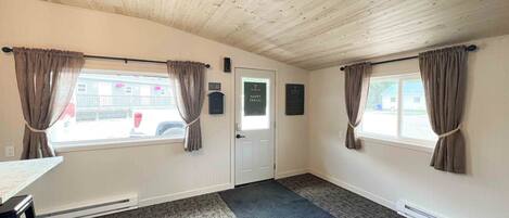 A room with wooden ceiling, two windows with curtains, a door, and a small table.