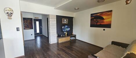 A spacious living area with a dark wood floor, a flat-screen TV mounted on a black wall, and a large painting of a sunset.