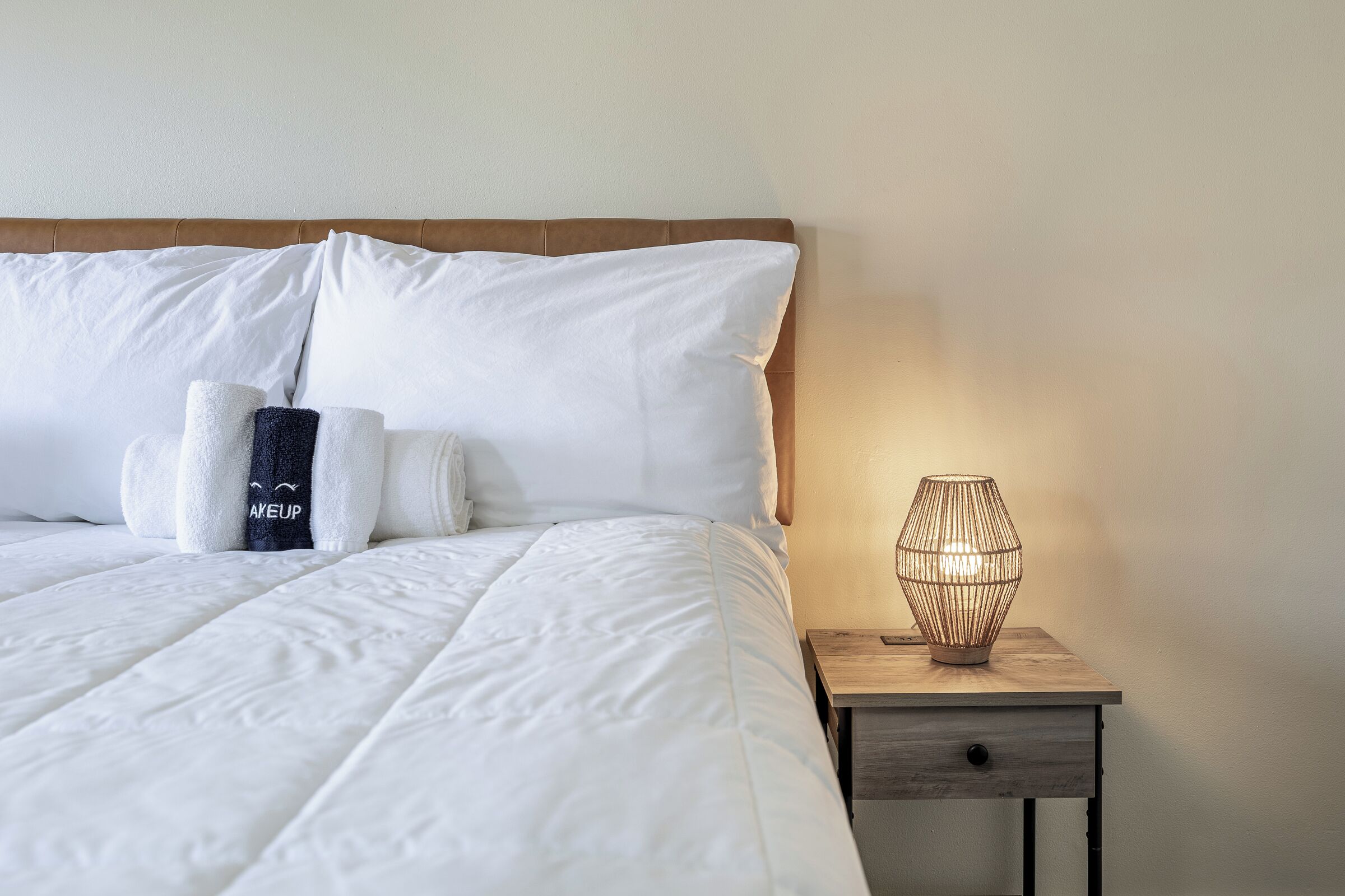 A neatly made bed with white bedding and pillows, a bedside table with a lamp, and a small drawer.