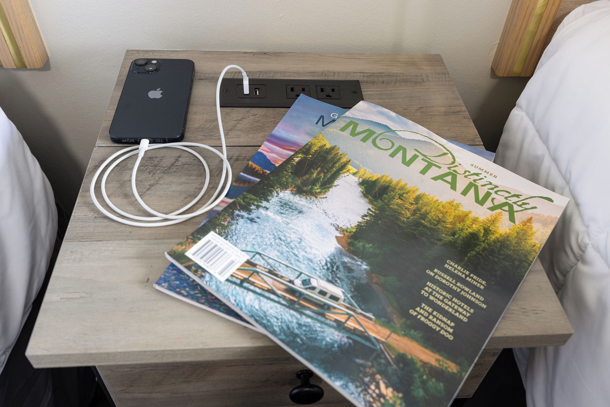 Une table de chevet en bois avec un téléphone intelligent, un chargeur et un magazine intitulé « Montana ».
