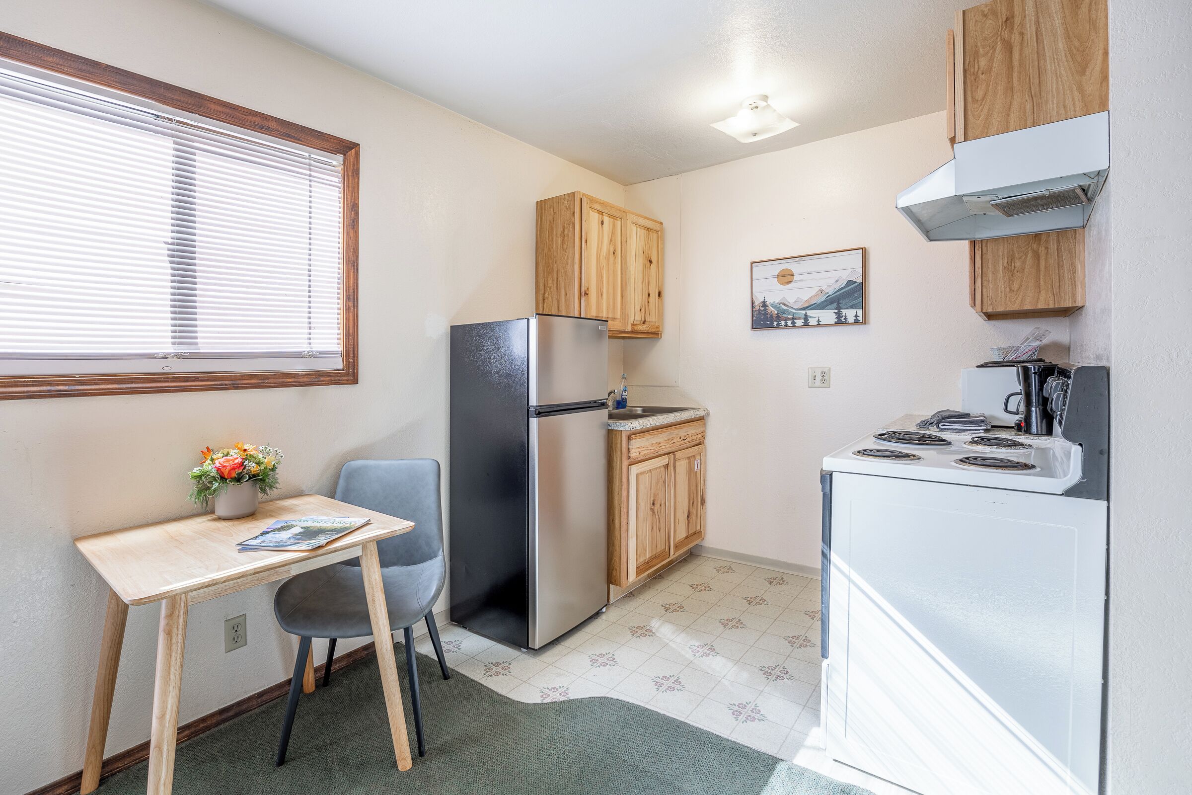 Une cuisine compacte comprenant un réfrigérateur, une cuisinière, des placards en bois, une petite table avec une chaise et une fenêtre avec des stores.
