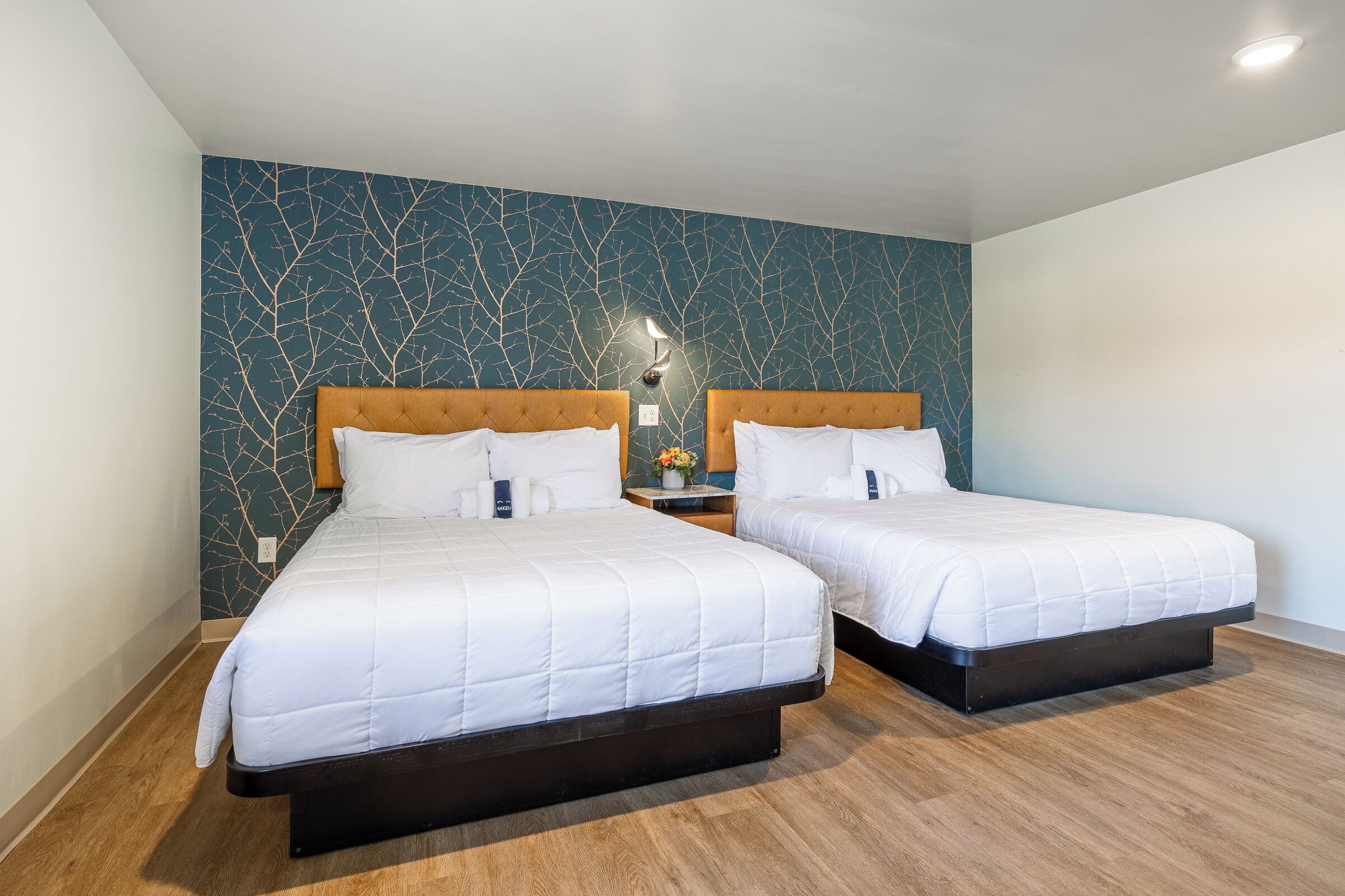 Une chambre d’hôtel avec deux lits, une table de chevet avec une lampe et un mur décoré d’un motif représentant une branche d’arbre.