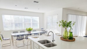 A modern kitchen with a central island, white cabinetry, and a dishwasher. There are two windows with blinds, a dining area with chairs, and a potted plant on the island. - Lovely Villa at Kissimmee (Kissimmee)