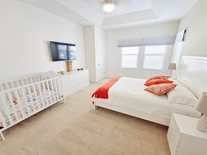 A bedroom with a white crib, a large bed with orange pillows, a TV on the wall, and a window with blinds. - Lovely Villa at Kissimmee (Kissimmee)