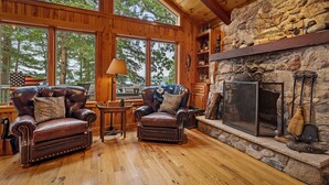 A cozy cabin interior with leather armchairs, a stone fireplace, and large windows offering a view of the forest.