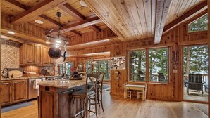 A wooden cabin interior with a kitchen island, bar stools, and large windows offering views of the outdoors.