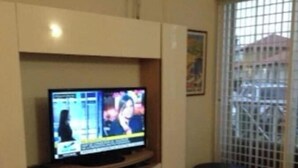 Uma sala de estar com uma TV de tela plana em um suporte, um sofá e uma janela com grade de metal.