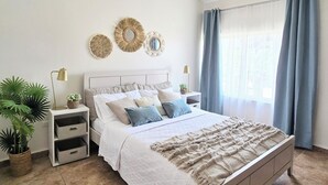 A bedroom with a bed, bedside tables, lamps, decorative wall mirrors, and potted plants.