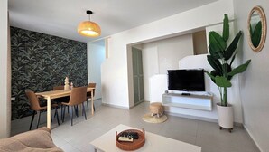 A modern living room with a dining area, a flat-screen TV, a large potted plant, and a patterned wallpaper.