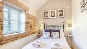 Un dormitorio con pared de piedra, una cama con ropa blanca, dos lámparas de noche y tres cuadros en la pared.