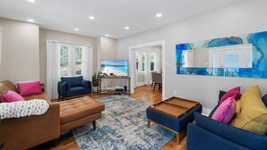 Una sala moderna con un sofá marrón, sillones azules y una mesa de centro de madera. Hay un televisor sobre un mueble bajo, un cuadro en la pared y vistas al exterior a través de amplias ventanas.