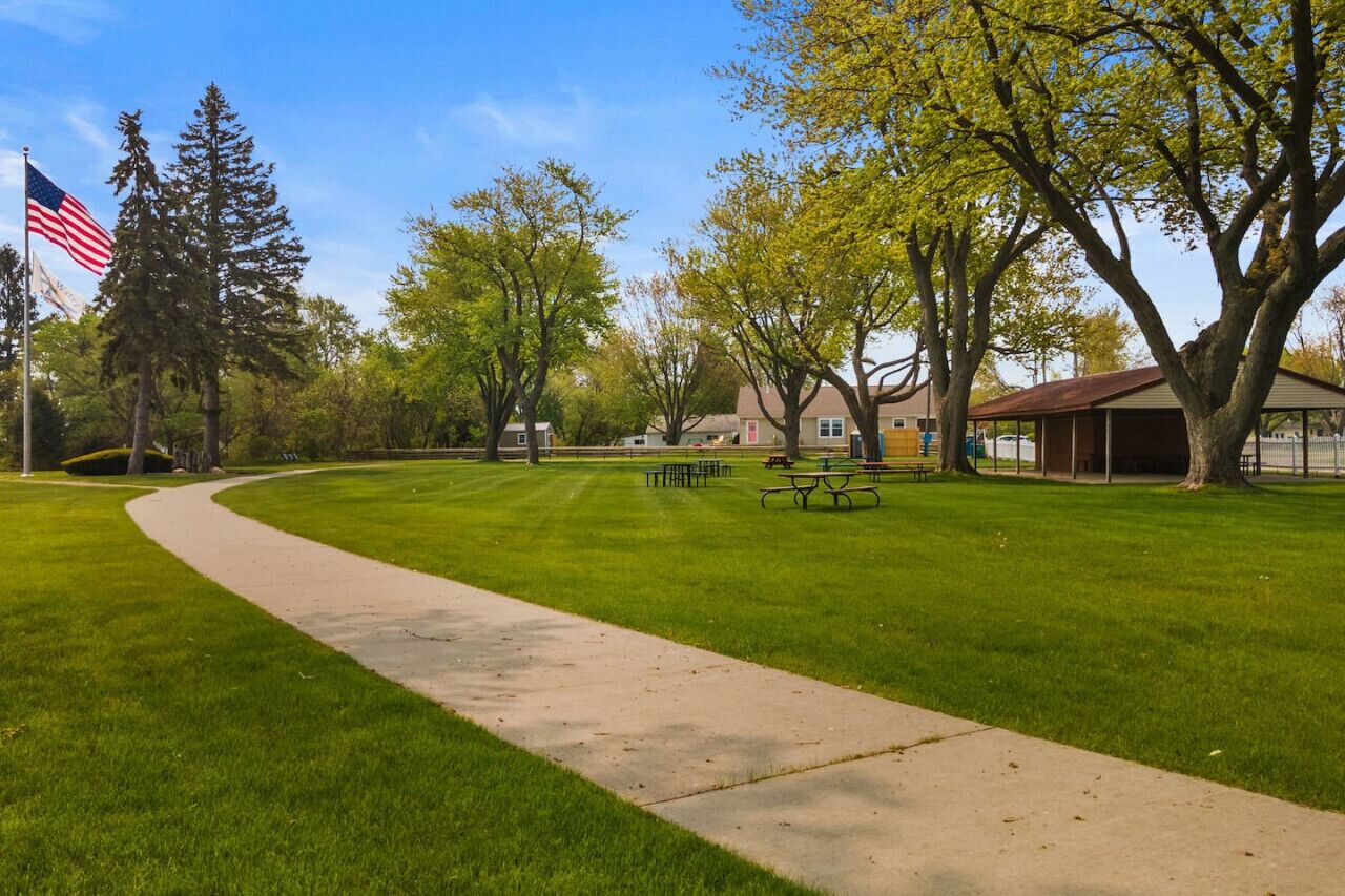 A park with a paved walkway, picnic tables, and a flagpole with an American flag.