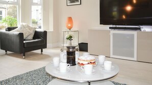 A modern living room with a sofa, a coffee table with cups and a cake, a TV, and a small cabinet. - 3 bedroom accommodation in Alnwick (Alnwick)
