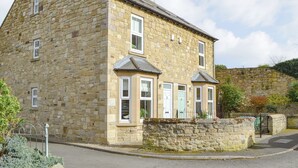A stone building with a gabled roof, multiple windows, and a stone wall in the background. - 3 bedroom accommodation in Alnwick (Alnwick)