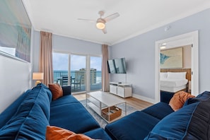 A modern living room with a sofa set, a flat-screen TV, a ceiling fan, and a balcony with a view. - Coastal Condo with Gulf Facing Views | Sleeps 7 (Gulf Shores)
