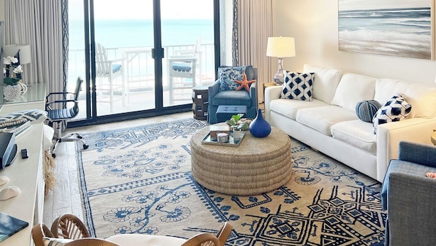 Une chambre d’hôtel avec un balcon, un canapé blanc, un fauteuil bleu, un pouf rond et un grand tableau représentant l’océan.