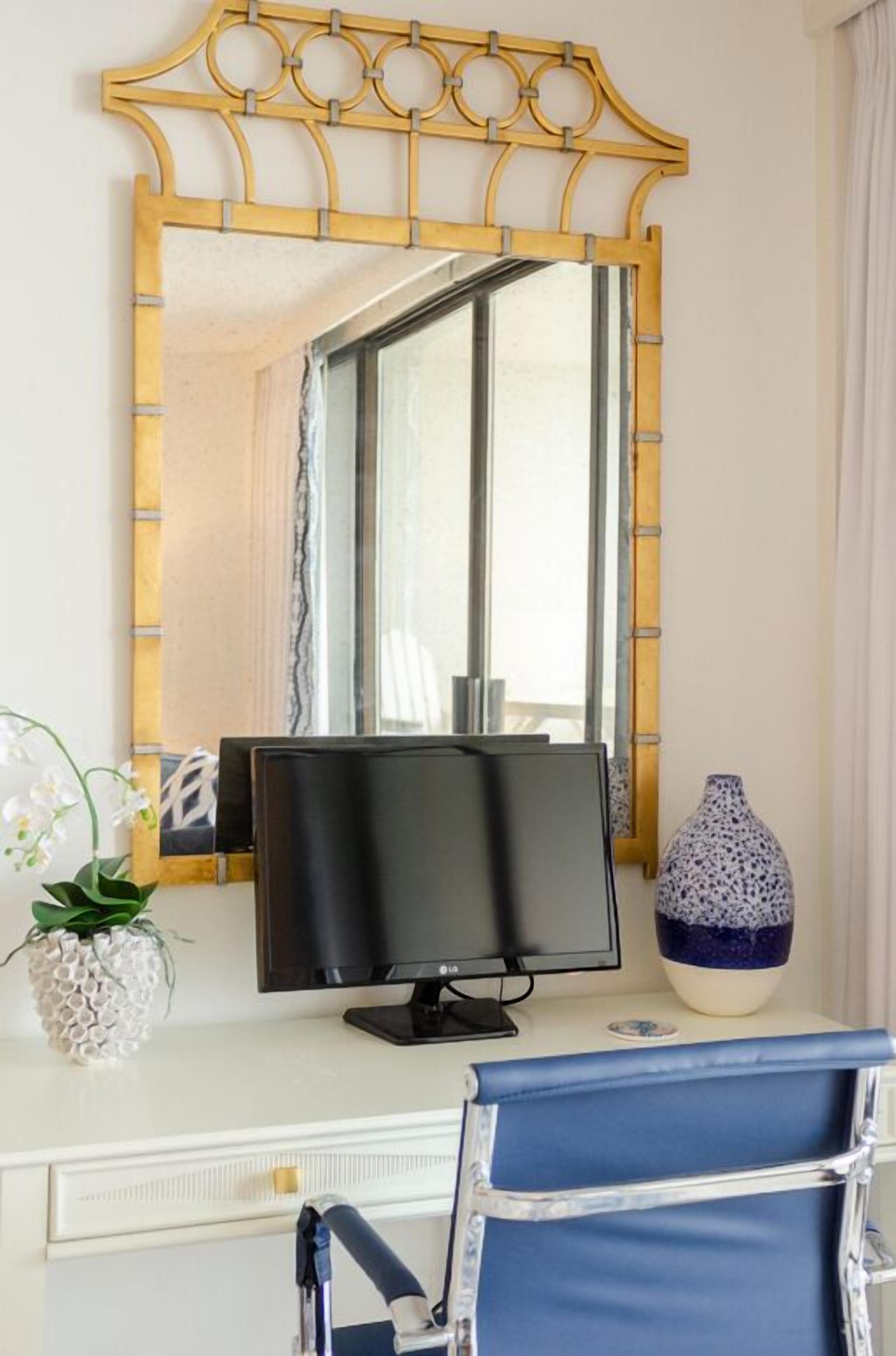 Una habitación con un escritorio blanco, un monitor negro, una silla azul, un espejo decorativo y un jarrón blanco.