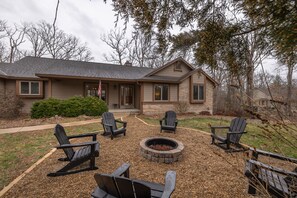 Une maison avec un foyer extérieur et quatre chaises Adirondack dans le jardin.