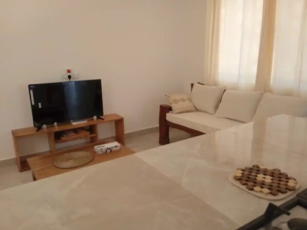 Ein Wohnzimmer mit einem Flachbildfernseher auf einem Holzständer, einem beigen Sofa und einem Couchtisch mit einem Gesellschaftsspiel.
