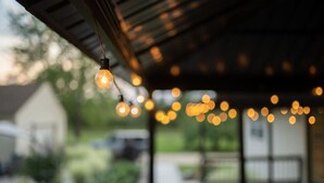 Un espacio al aire libre cubierto, con luces colgantes y un fondo desenfocado de vegetación y un edificio.