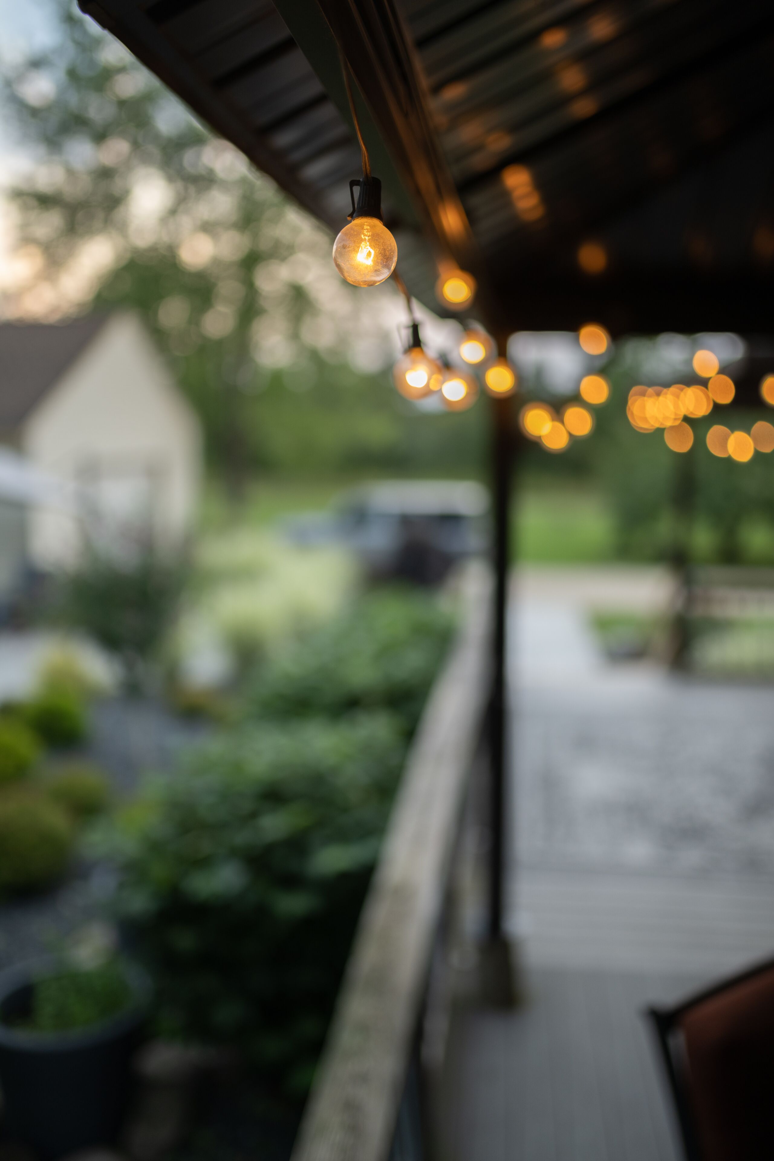 A porch with hanging lights and a view of a garden and house in the background.