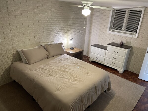 Un dormitorio con cama, mesita de noche, cómoda, ventana con persianas y ventilador de techo.