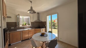 A compact kitchen with wooden cabinets, a round dining table, and a view of the garden through a sliding door.