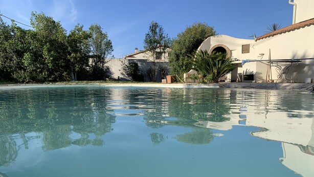A clear pool with reflections of trees and buildings, surrounded by greenery and a white building with a tiled roof.
