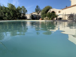 Une piscine aux eaux limpides où se reflètent les arbres et les bâtiments, entourée de verdure et bordée d’un bâtiment blanc au toit recouvert de tuiles.