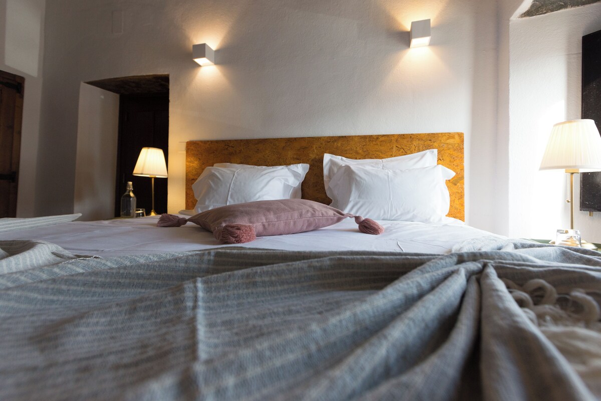 A hotel room with a bed, pillows, a headboard, and wall-mounted lamps.