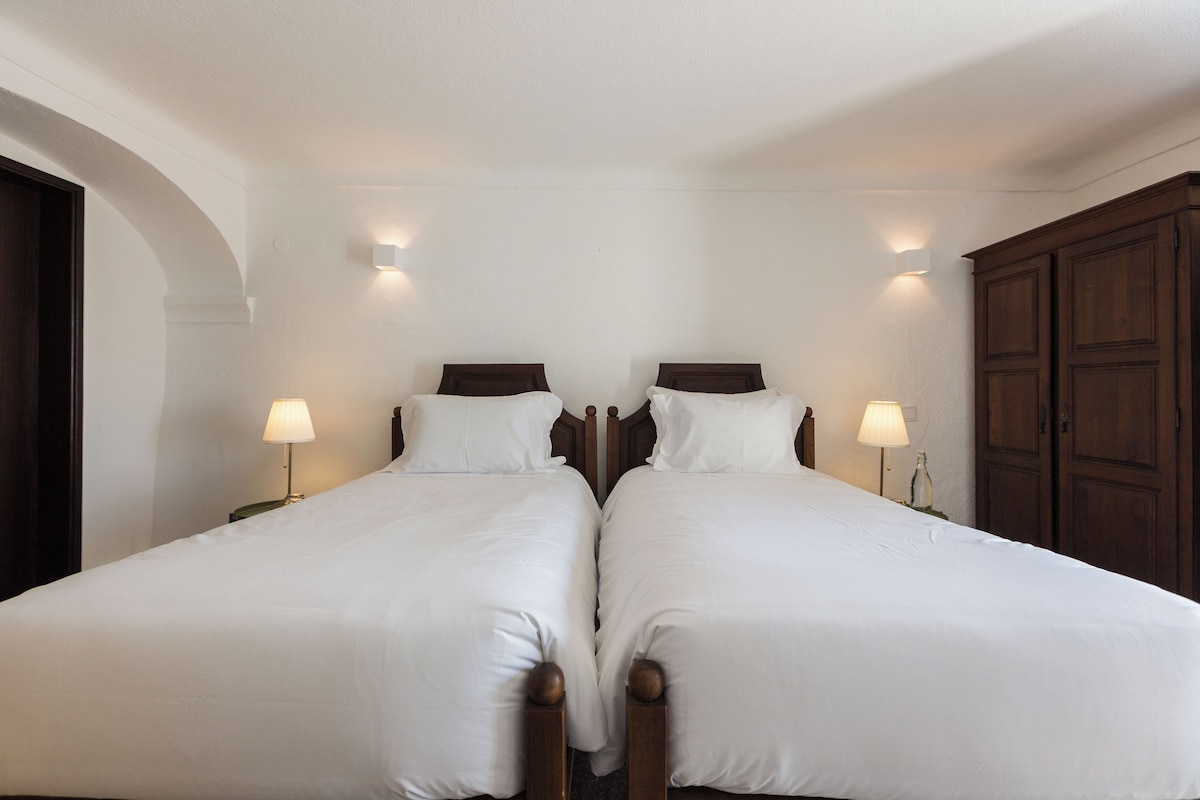 A double bed with white linens and wooden headboards, two bedside lamps, and a wooden wardrobe.