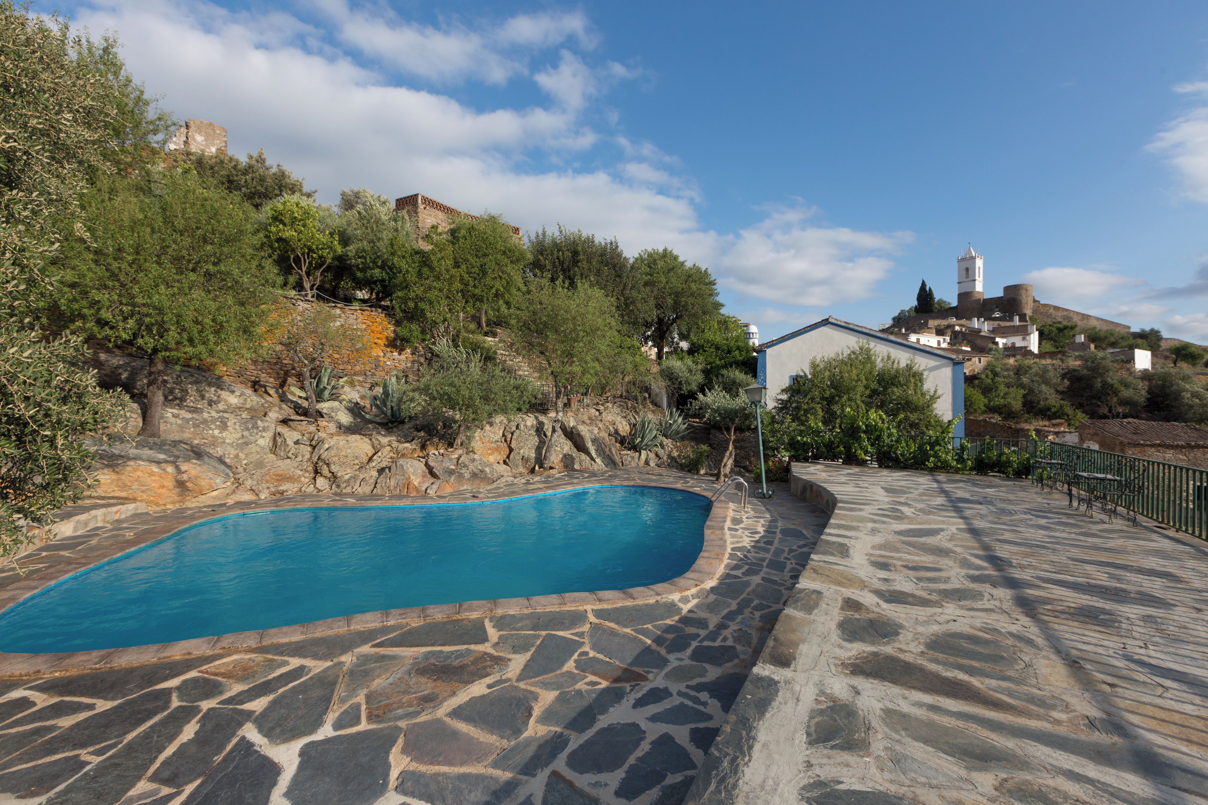 Une piscine en pierre avec une terrasse en pierre, entourée de verdure et bordée d’une colline où se dressent des bâtiments en arrière-plan.