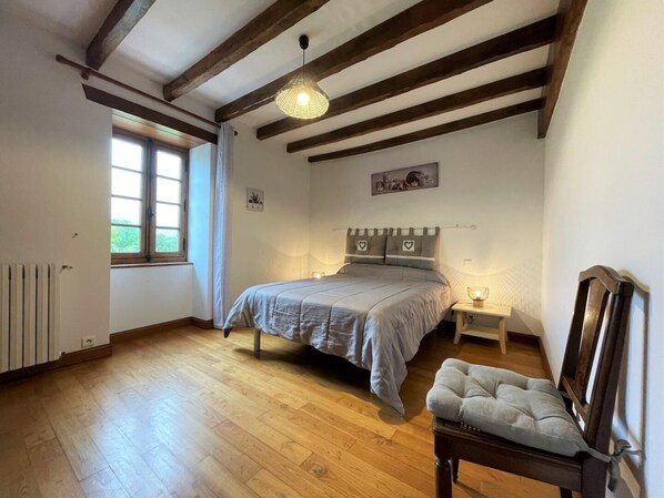 A bedroom with a wooden floor, a bed with grey bedding, a bedside table with a lamp, a chair, and a window with curtains. - Zamatelua 1 - Sare - Gîte (SARE)