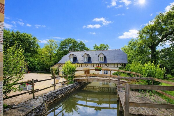 A traditional half-timbered house with a stone bridge over a pond, surrounded by greenery.