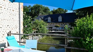 A wooden deck with a table and chairs, a stone wall, and a house with a dark roof and white windows in the background. - LE Mouly/petit Gite - Miossens-lanusse (MIOSSENS-LANUSSE)