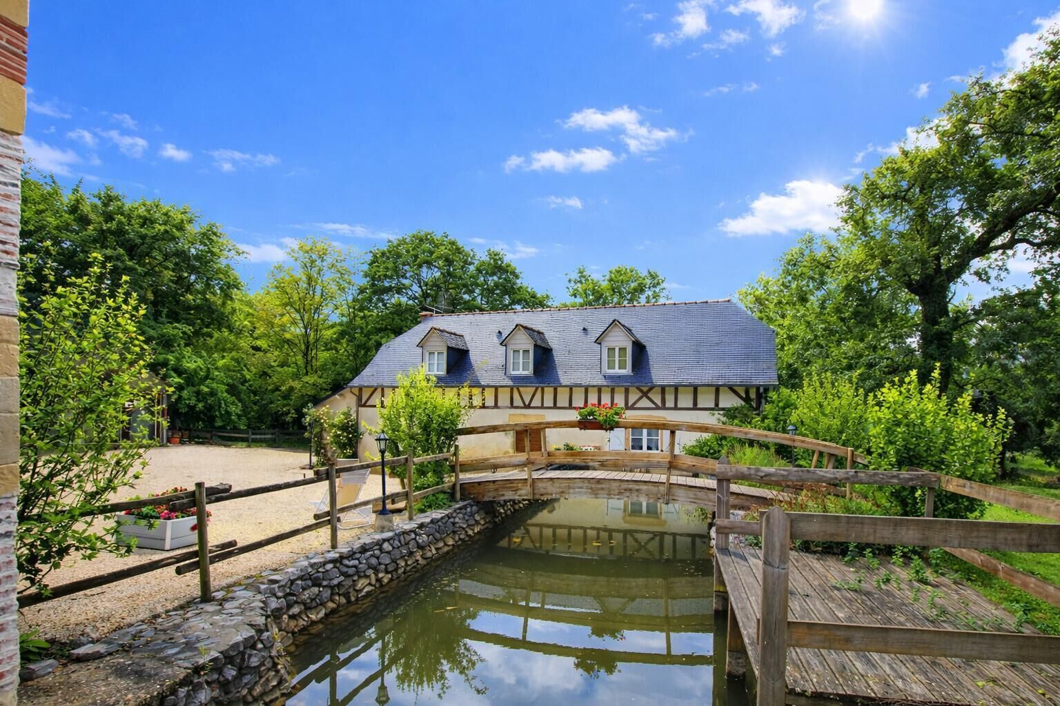 Uma casa tradicional de enxaimel com uma ponte de pedra sobre um lago, cercada por vegetação.