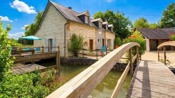 Een rustiek stenen gebouw met een houten terras boven een waterkanaal, omgeven door groen.
