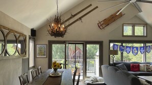 A dining area with a long wooden table, chairs, and a chandelier. There is a gray sofa and a dog bed in the living area. - Crested Butte Home | 4 Bedroom | Hot Tub | Walk to Elk | Gas Grill |Pet-Friendly (Crested Butte)