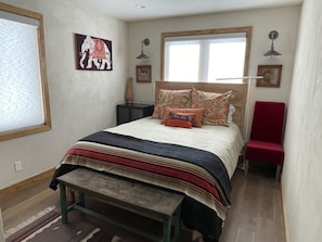 A bedroom with a bed, a red chair, a nightstand, a lamp, and a window.