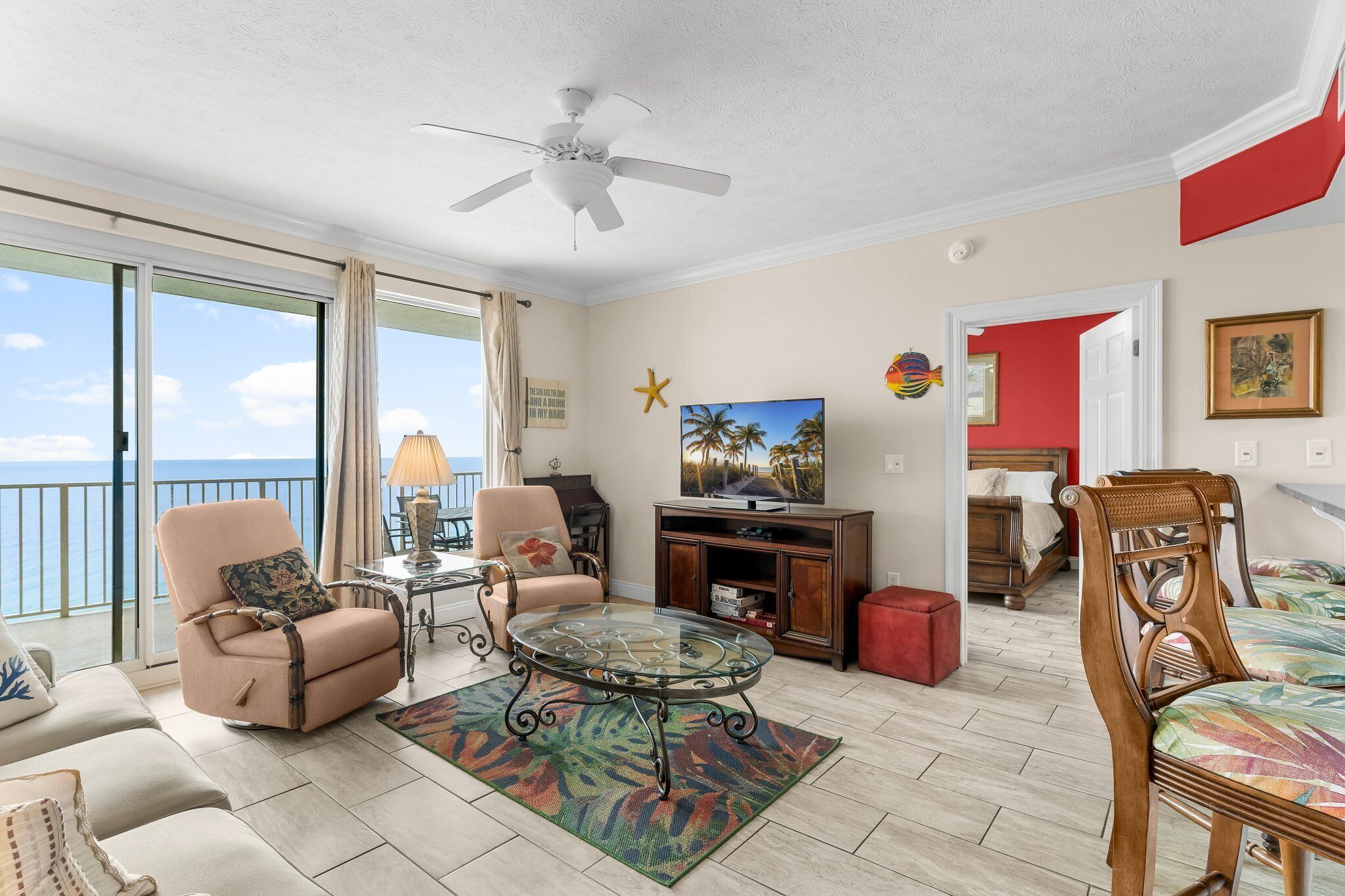 A living room with a glass coffee table, beige armchairs, a wooden chair, a television, and a view of the ocean.