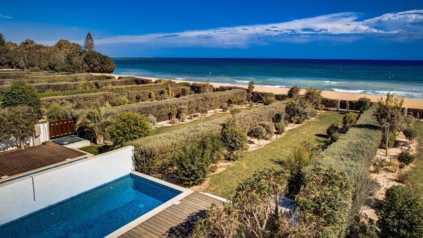 A pool with a wooden deck, surrounded by well-maintained gardens and a view of the ocean. - Atlantis Gardens Resort (Mazotos)