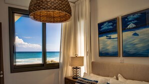 A bedroom with a large window overlooking the sea, a bed with patterned pillows, a nightstand with a lamp, and two framed paintings of boats on the water. - Atlantis Gardens Resort (Mazotos)