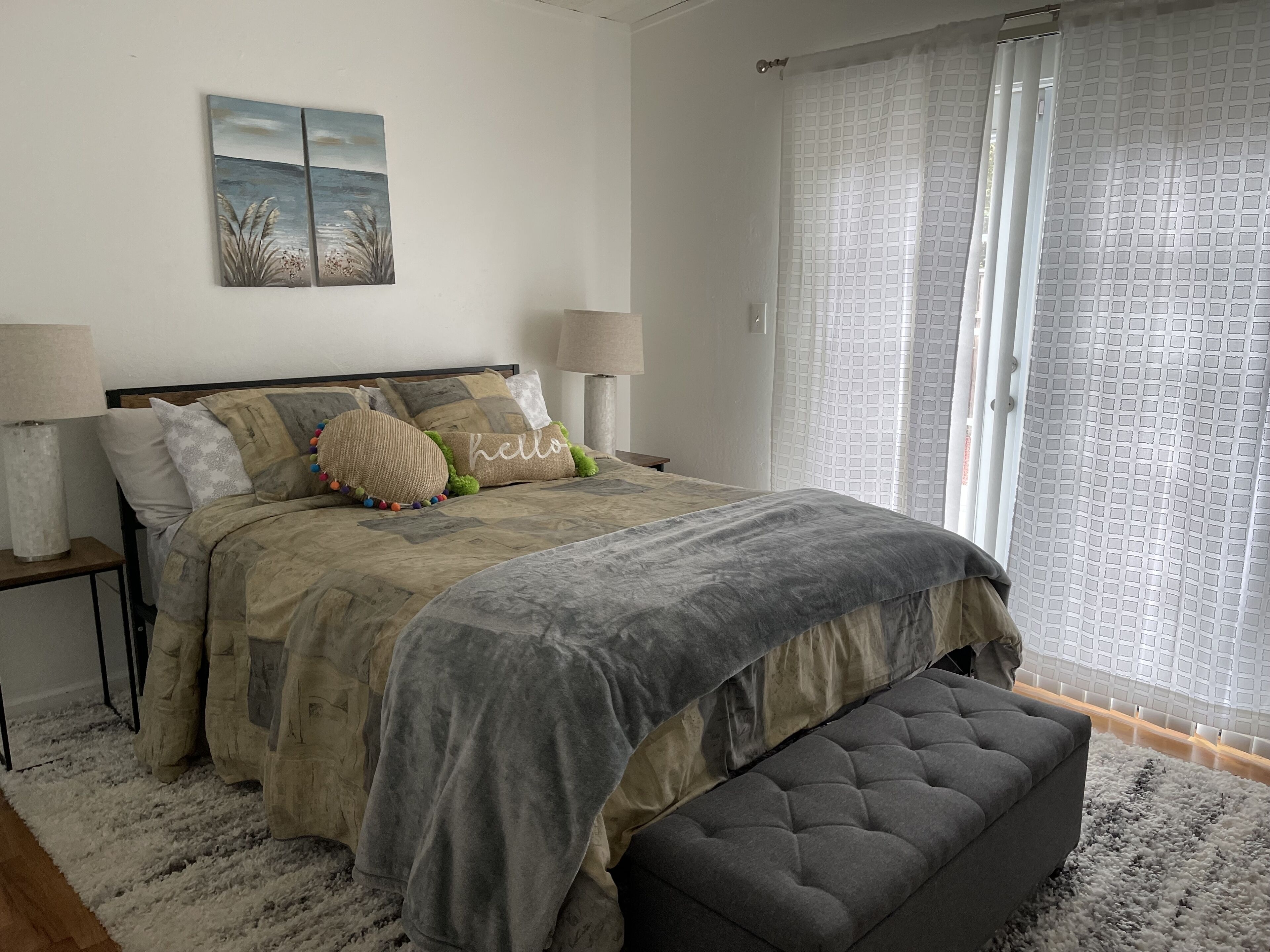 Een slaapkamer met een bed, twee nachtkastjes, een bankje, een aan de muur hangend schilderij en een raam met een gordijn met patroon.