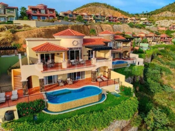 Une maison à plusieurs étages dotée d’une grande piscine, de balcons et d’un paysage verdoyant luxuriant.
