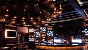 A bar area with a curved counter, multiple seating options, and a modern ceiling design with exposed beams and lighting fixtures.