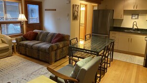Una sala de estar acogedora con un sofá, una mesa de comedor con superficie de vidrio y una zona de cocina con armarios de madera y un refrigerador.