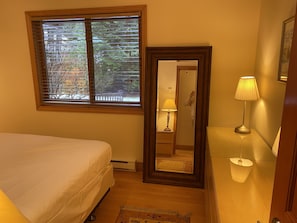 Un dormitorio con cama, ventana con persianas, espejo, lámpara de noche y alfombra.