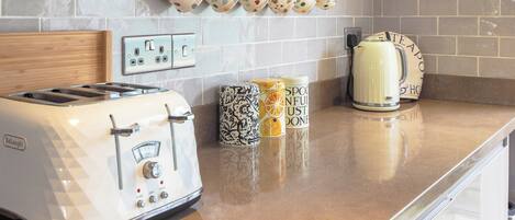 A kitchen with a white De'Longhi toaster, a kettle, and a coffee maker on the counter. There are colorful mugs hanging on the wall and a spice rack below.
